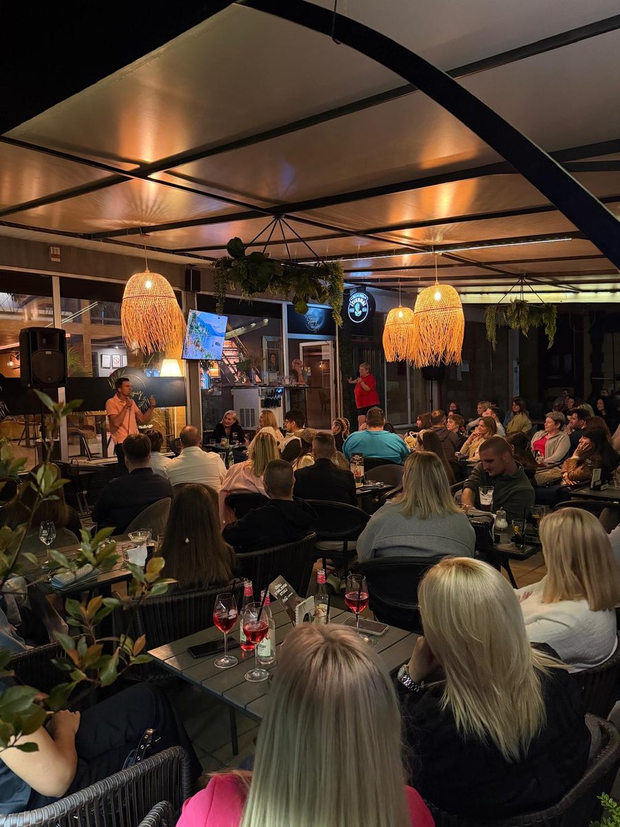 Puna bašta publike na stand-up večeri pod pletenim lampama